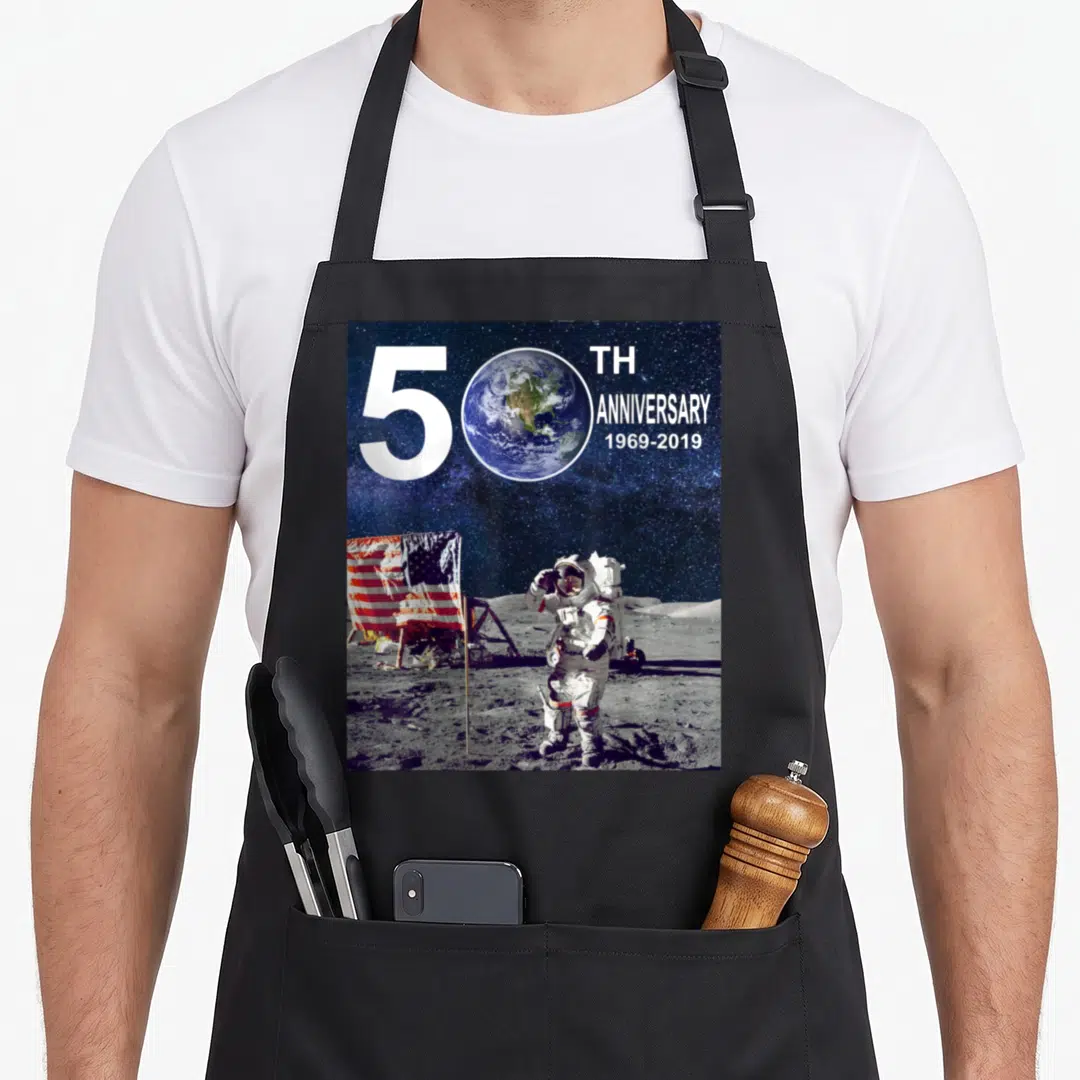 50th anniversary moon apollo apron - Astronaut and flag on moon surface with 50th anniversary 1969-2019 text and Earth — shown on a black cotton canvas apron against a white background
