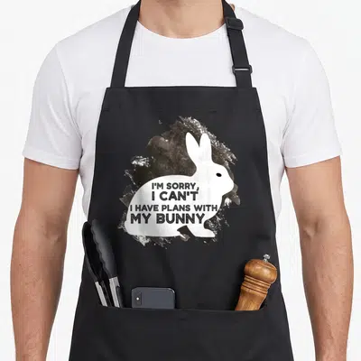 Sorry paint apron - White bunny silhouette with black text over brown and white paint splatter — shown on a black cotton canvas apron against a white background