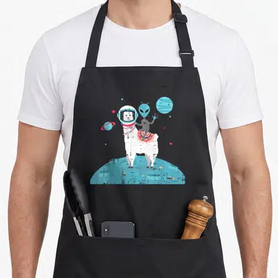 Space chef outfit with apron - White llama in astronaut helmet with blue alien rider on blue moon surface — shown on a black cotton canvas apron against a white background