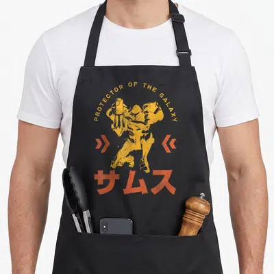 Samus nebula apron - Yellow armored character with PROTECTOR OF THE GALAXY text and orange Japanese characters — shown on a black cotton canvas apron against a white background