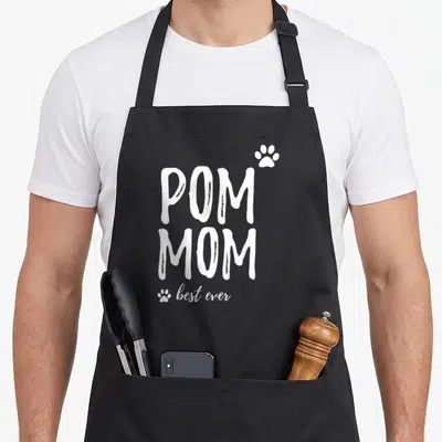 Funny pom dog apron - White stacked text reading POM MOM best ever with two paw prints on black fabric — shown on a black cotton canvas apron against a white background