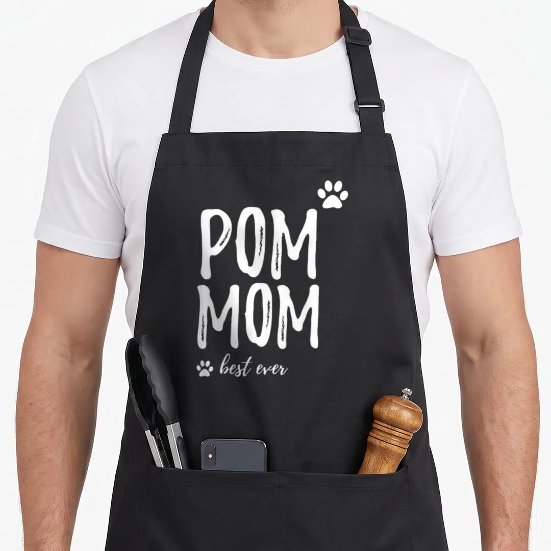 Funny pom dog apron - White stacked text reading POM MOM best ever with two paw prints on black fabric — shown on a black cotton canvas apron against a white background