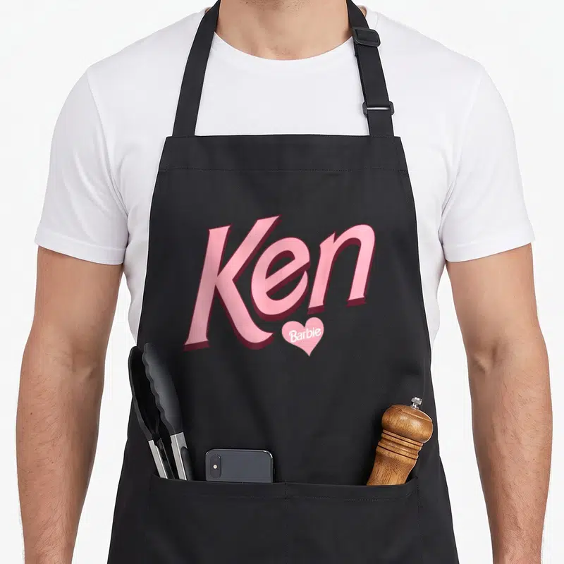 Ken valentine apron - Large pink Ken text with small pink Barbie heart below on white background — shown on a black cotton canvas apron against a white background