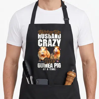 Pig husband wife aprons - Two realistic guinea pigs in white orange brown with Driving My Husband Crazy text — shown on a black cotton canvas apron against a white background