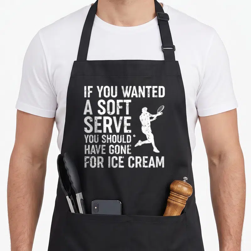 Wanted to serve man apron - White distressed text stacking beside a tennis player silhouette serving a ball — shown on a black cotton canvas apron against a white background