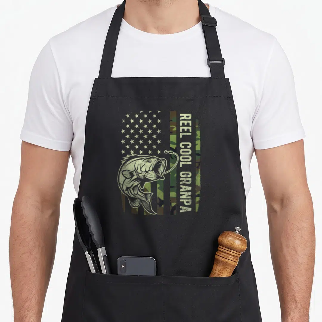 Reel grandpa american flag apron - Distressed bass fish over camo flag with vertical REEL COOL GRANPA text in yellow — shown on a black cotton canvas apron against a white background