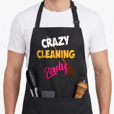 Cleaning housekeeper apron - Stacked Crazy Cleaning Lady text with diagonal cartoon broom in white yellow pink and brown — shown on a black cotton canvas apron against a white background