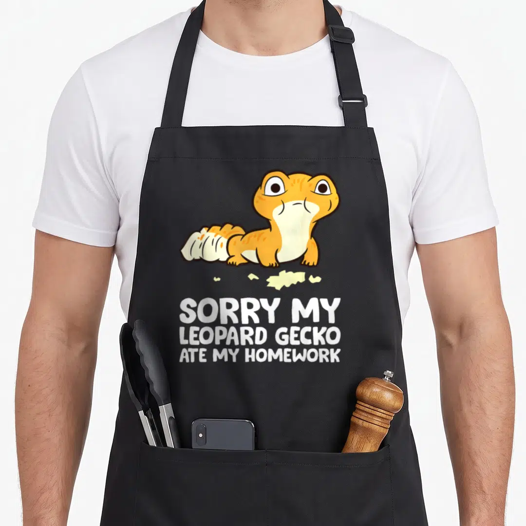 Sorry leopard apron - Cartoon orange leopard gecko with yellow paper scraps above white text reading sorry my leopard — shown on a black cotton canvas apron against a white background