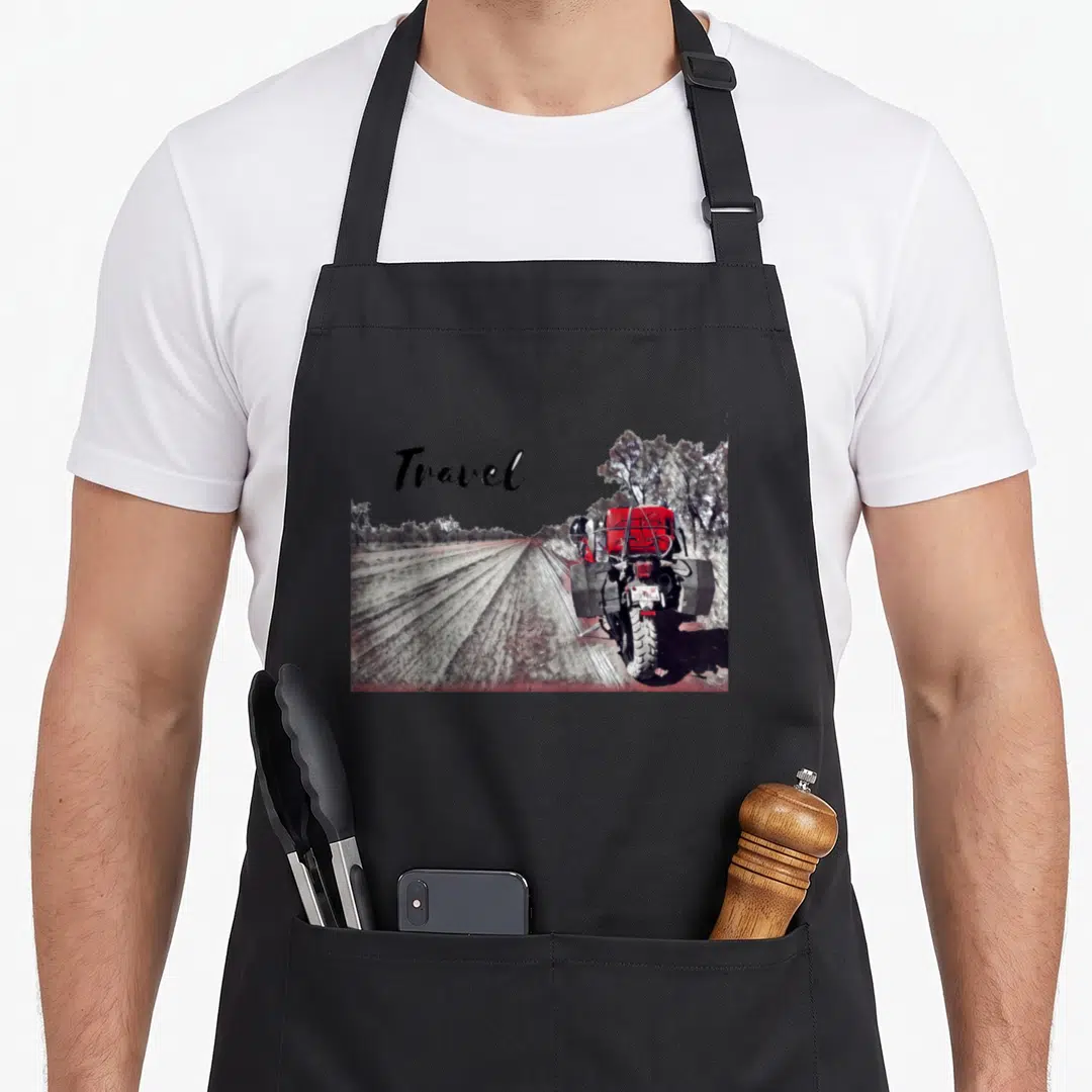 Travel bike apron - Word Travel above photo of motorcycle on dirt road with red tank and grey trees — shown on a black cotton canvas apron against a white background