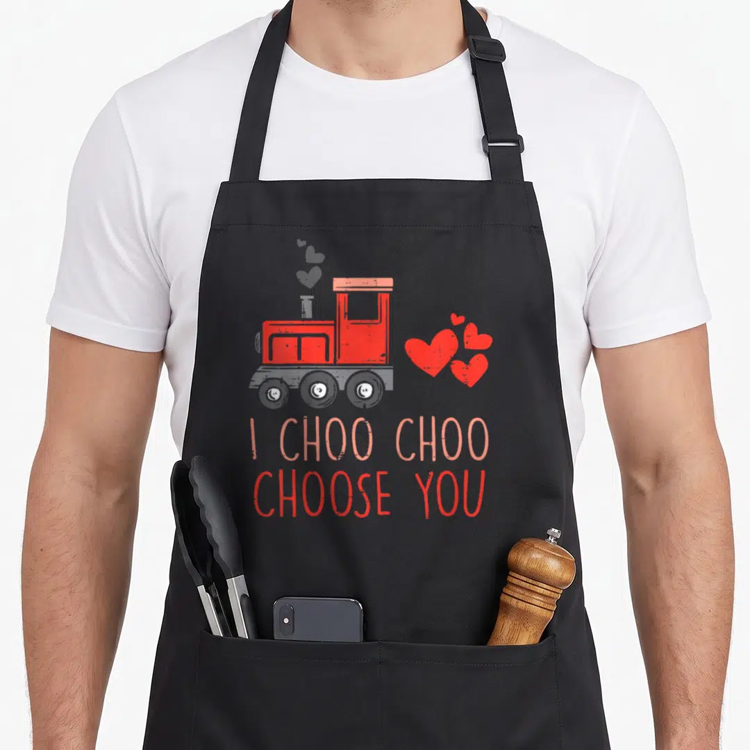 Choo valentine's day apron - Red toy train with grey and pink hearts and text I CHOO CHOO CHOOSE YOU — shown on a black cotton canvas apron against a white background