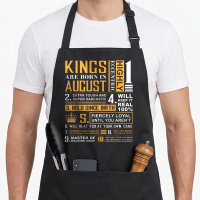 Birthday august king apron - Orange and white text grid listing August birth traits including fierce loyalty and sarcasm — shown on a black cotton canvas apron against a white background