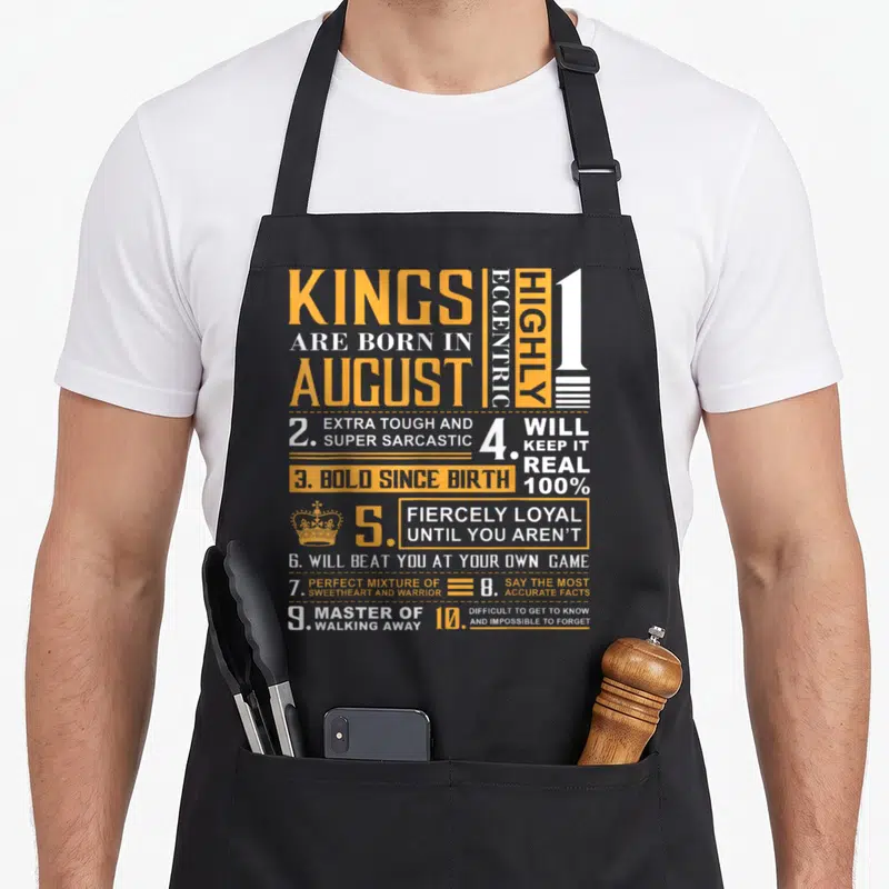 Birthday august king apron - Orange and white text grid listing August birth traits including fierce loyalty and sarcasm — shown on a black cotton canvas apron against a white background