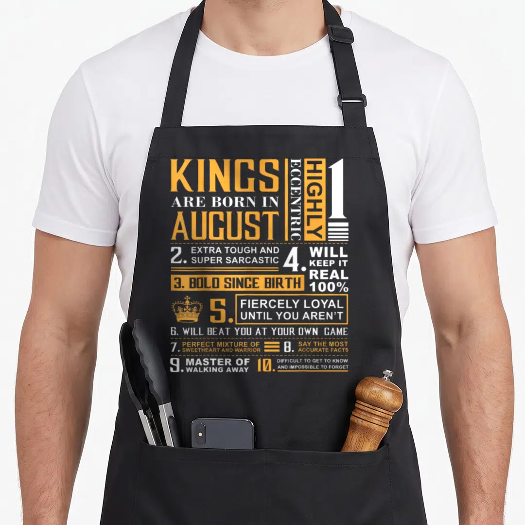 Birthday august king apron - Orange and white text grid listing August birth traits including fierce loyalty and sarcasm — shown on a black cotton canvas apron against a white background