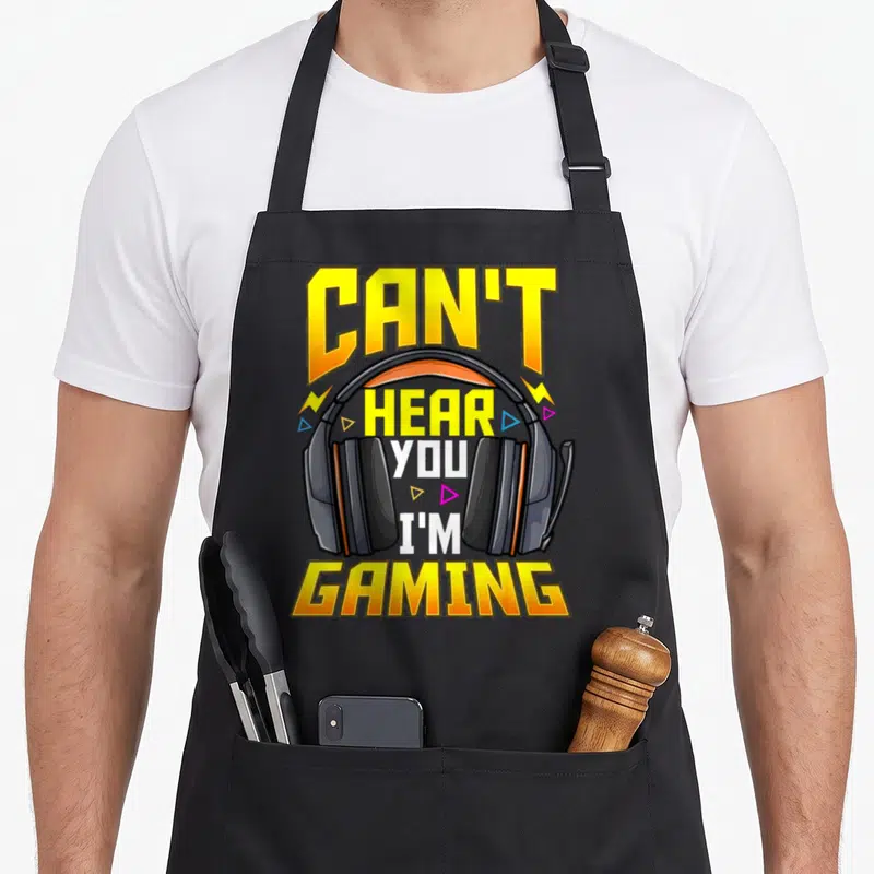 Cute hear gaming apron - Cartoon headset with yellow CAN'T HEAR YOU I'M GAMING text and lightning bolts — shown on a black cotton canvas apron against a white background