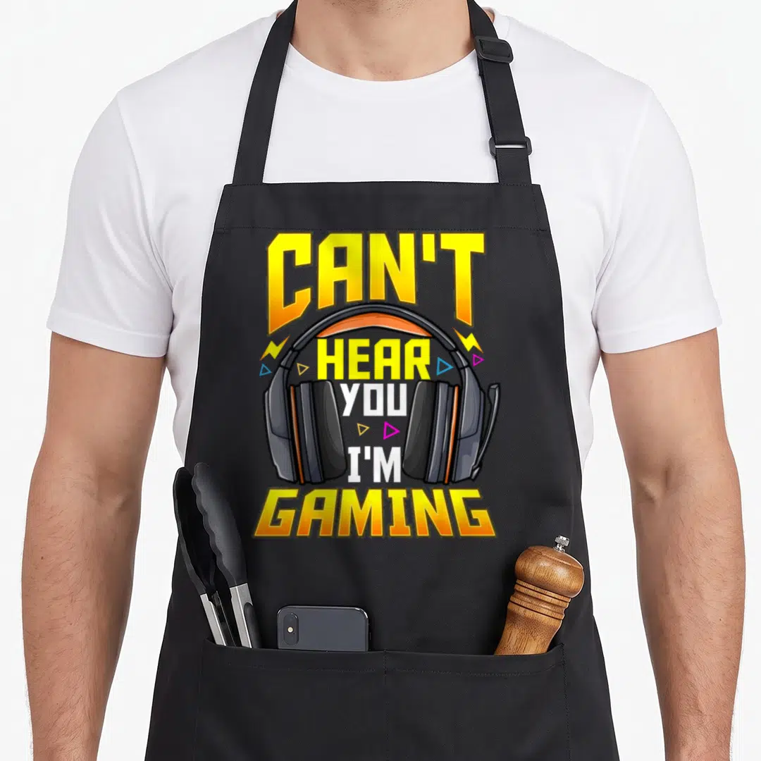 Cute hear gaming apron - Cartoon headset with yellow CAN'T HEAR YOU I'M GAMING text and lightning bolts — shown on a black cotton canvas apron against a white background