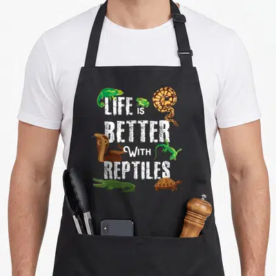Better pet apron - LIFE IS BETTER WITH REPTILES text surrounded by cartoon chameleon, python, cobra, lizard, alligator — shown on a black cotton canvas apron against a white background