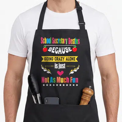 School friends apron - Colorful stacked text reading School Secretary Besties with apples, pencils, heart, and stars — shown on a black cotton canvas apron against a white background