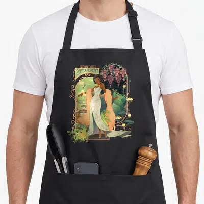 Tiana frog aprons - Cartoon woman in white dress with LIVIN THE BAYOU DREAM banner, purple wisteria, and alligator — shown on a black cotton canvas apron against a white background