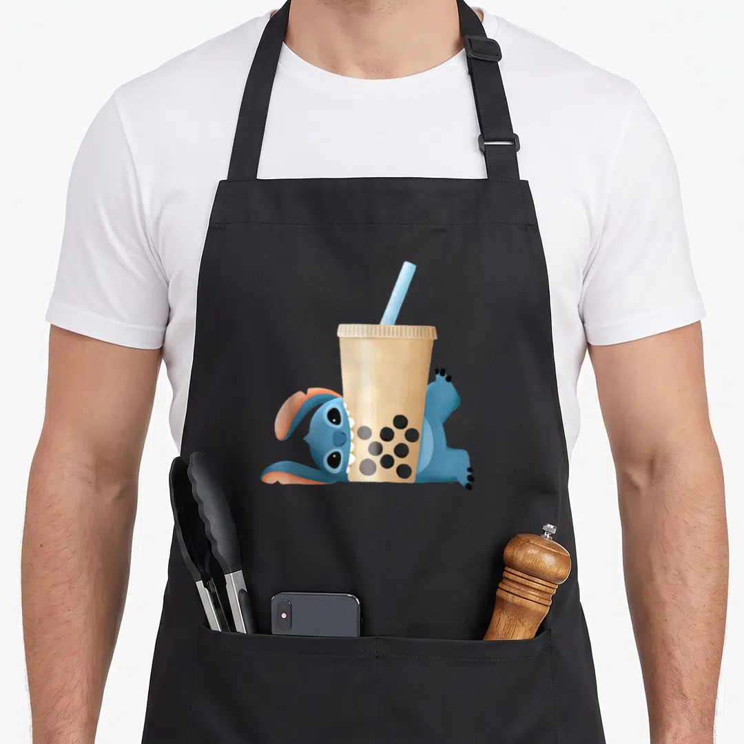 Aanhpi stitch apron - Blue alien lying flat behind beige cup with black tapioca pearls and straw — shown on a black cotton canvas apron against a white background