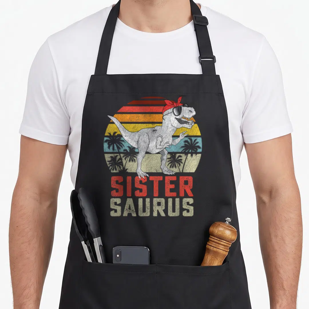 Sistersaurus t-rex apron - T-Rex in red bandana and sunglasses with stacked sister saurus text over striped sun — shown on a black cotton canvas apron against a white background