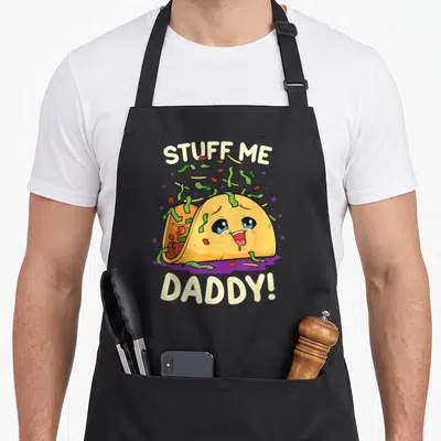 Game the dirty apron - Cartoon taco with face, falling green toppings, and arched text STUFF ME DADDY! — shown on a black cotton canvas apron against a white background