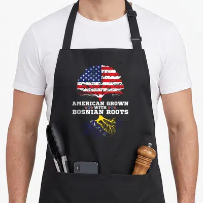 Grown bosnian girl apron - Tree with US flag canopy and yellow blue roots reading AMERICAN GROWN WITH BOSNIAN ROOTS — shown on a black cotton canvas apron against a white background