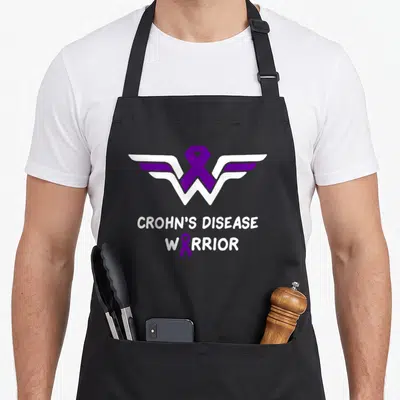 Crohn disease ribbon apron - Purple ribbon winged W logo with white text CROHN'S DISEASE WARRIOR — shown on a black cotton canvas apron against a white background