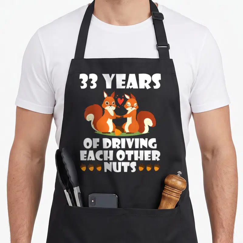 Fox husband driving aprons - Two orange squirrels holding hands with 33 years of driving each other nuts text and acorns — shown on a black cotton canvas apron against a white background