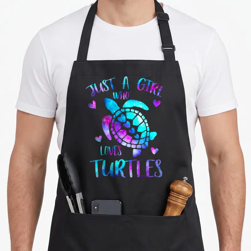 Turtle galaxies apron - Cartoon turtle with blue purple pink galaxy texture and hearts, text Just A Girl Who Loves — shown on a black cotton canvas apron against a white background