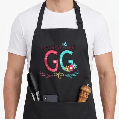 Colorful grandma apron - Blessed to be called GG text with pink G, blue G, flowers, and yellow swirl on black background — shown on a black cotton canvas apron against a white background