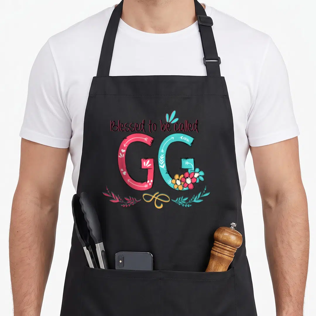 Colorful grandma apron - Blessed to be called GG text with pink G, blue G, flowers, and yellow swirl on black background — shown on a black cotton canvas apron against a white background