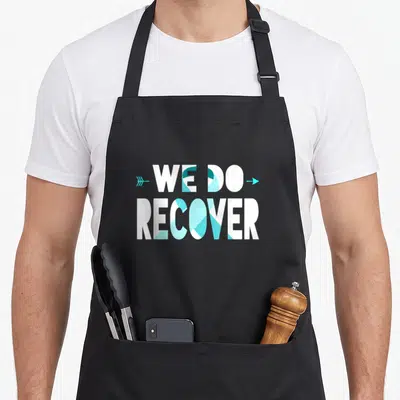 Recover ribbon apron - White text We Do Recover with teal arrow and blue geometric fills — shown on a black cotton canvas apron against a white background