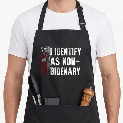 Non american flag apron - Rifle graphic with red stripes and white text I Identify As Non-Bidenary — shown on a black cotton canvas apron against a white background