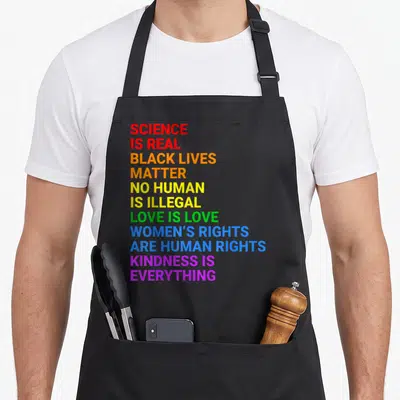 Rainbow gay pride apron - Stacked rainbow text reading Science Is Real Black Lives Matter Love Is Love — shown on a black cotton canvas apron against a white background