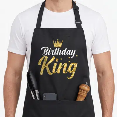 Son kings apron - Gold crown above white Birthday and glittery King text with small stars — shown on a black cotton canvas apron against a white background