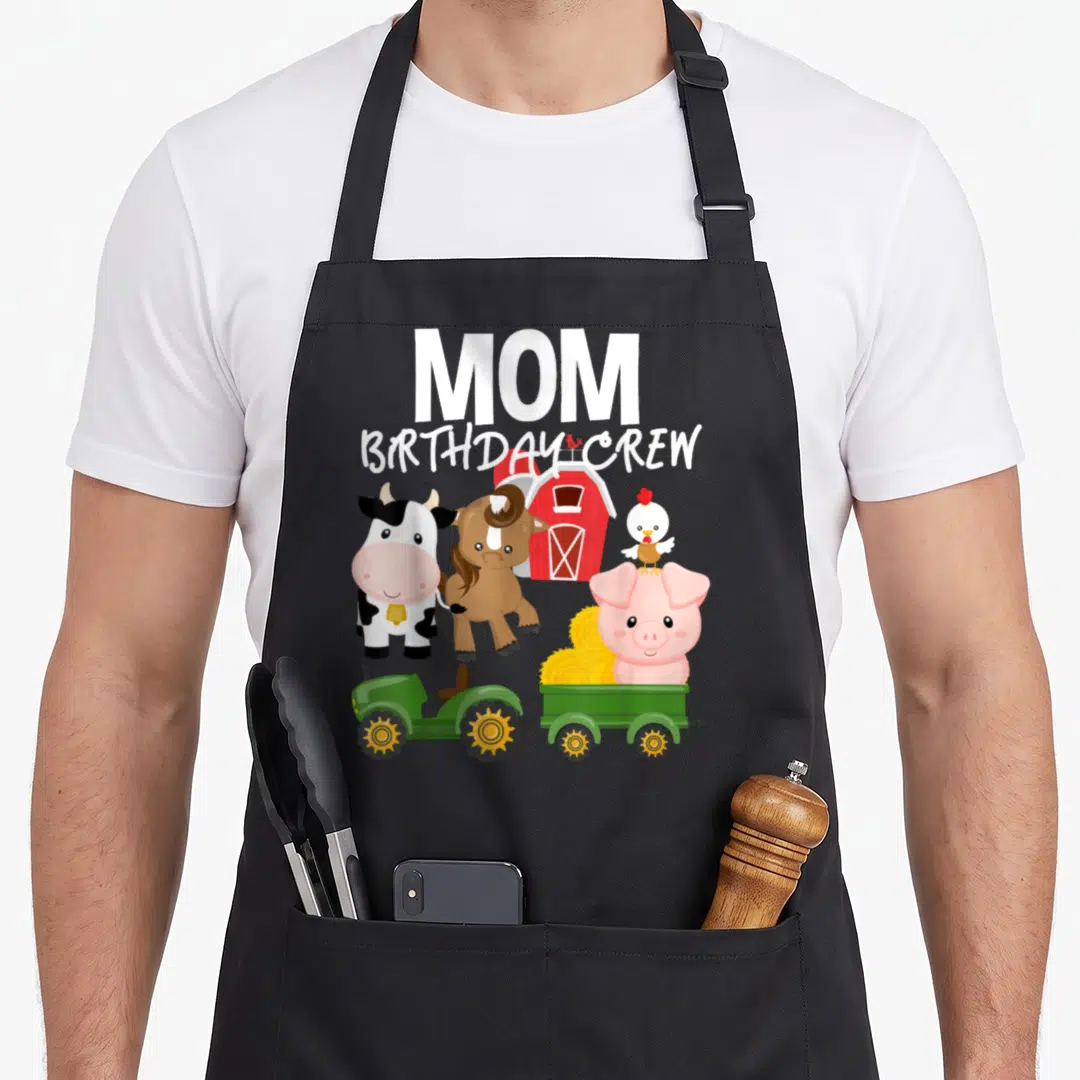 Farm crew aprons - Cartoon cow horse pig and green tractor with MOM BIRTHDAY CREW text — shown on a black cotton canvas apron against a white background