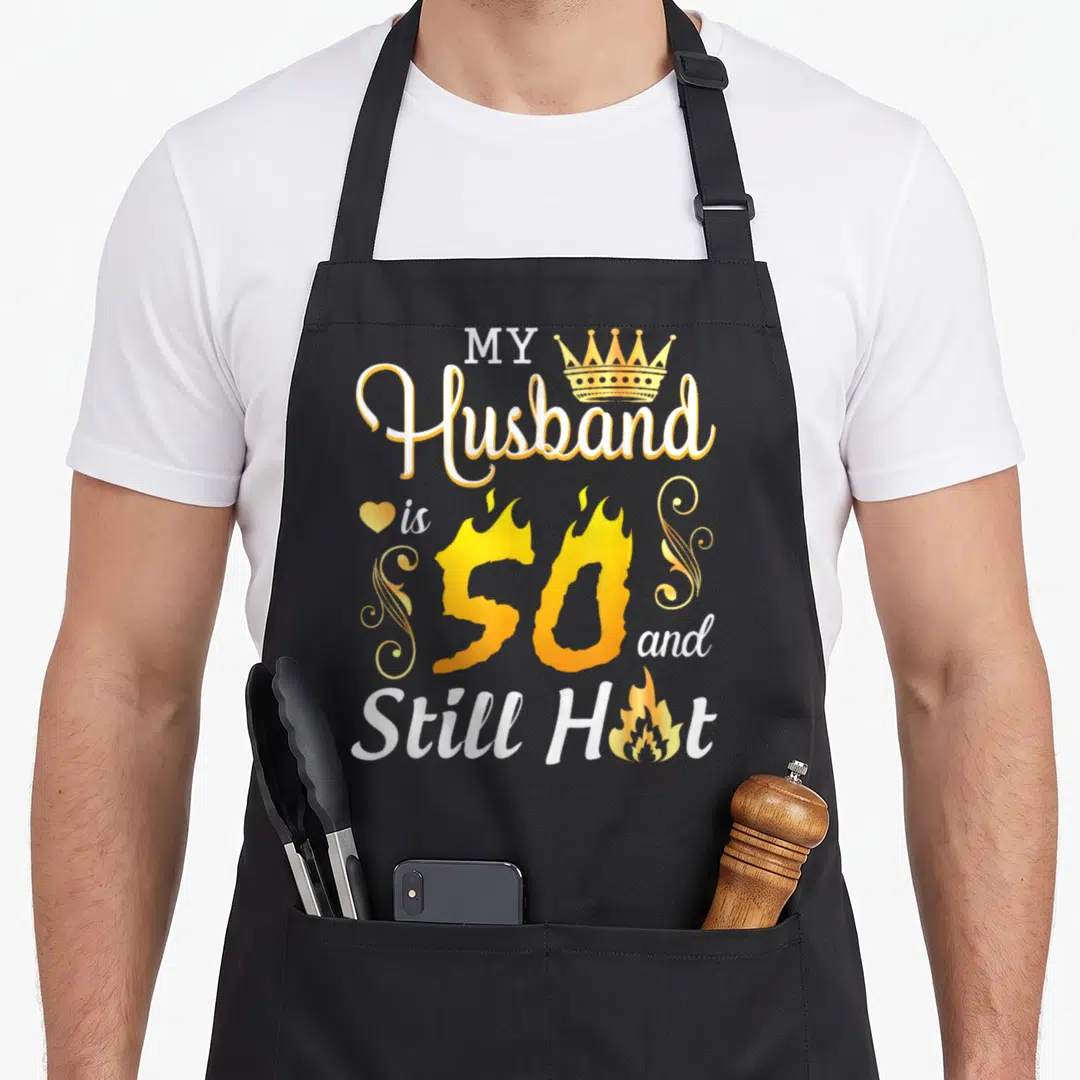 Hot husband wife aprons - My Husband is 50 and Still Hot text with flame fill and crown icon — shown on a black cotton canvas apron against a white background