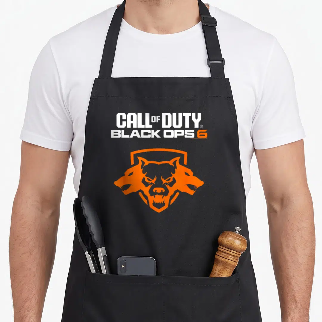 Ops video game apron - Stacked white and orange Call of Duty Black Ops 6 text above orange three-headed wolf shield — shown on a black cotton canvas apron against a white background
