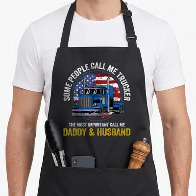 Trucker husband father day apron - Blue semi-truck over distressed USA flag with text about being a daddy and husband — shown on a black cotton canvas apron against a white background