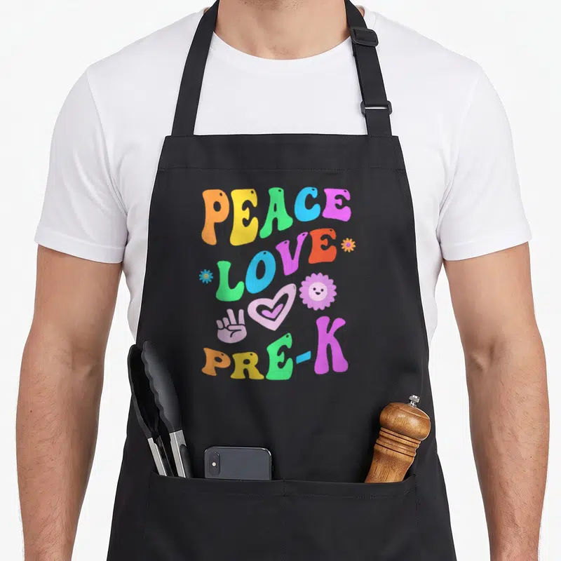 Miss teacher apron - Retro text PEACE LOVE PRE-K with peace sign, heart, and flowers in orange, yellow, green, blue — shown on a black cotton canvas apron against a white background