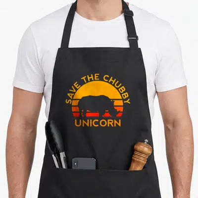 Chubby unicorn apron - Rhino silhouette against orange red striped sun with save the chubby unicorn text — shown on a black cotton canvas apron against a white background