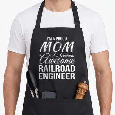 Railroad mother's day aprons - White stacked typography reading I'm a proud mom of a freaking awesome railroad engineer — shown on a black cotton canvas apron against a white background