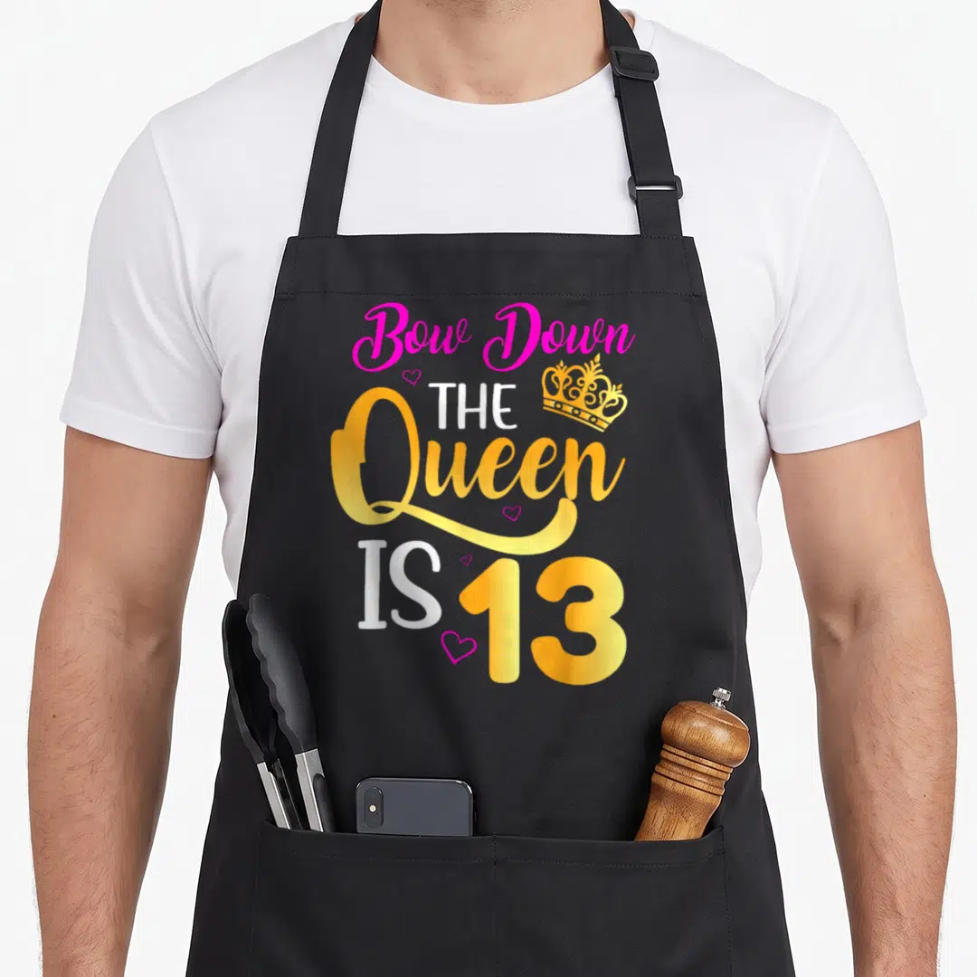 Funny 13th general birthday apron - Stacked pink yellow and white text reading Bow Down The Queen Is 13 with crown — shown on a black cotton canvas apron against a white background