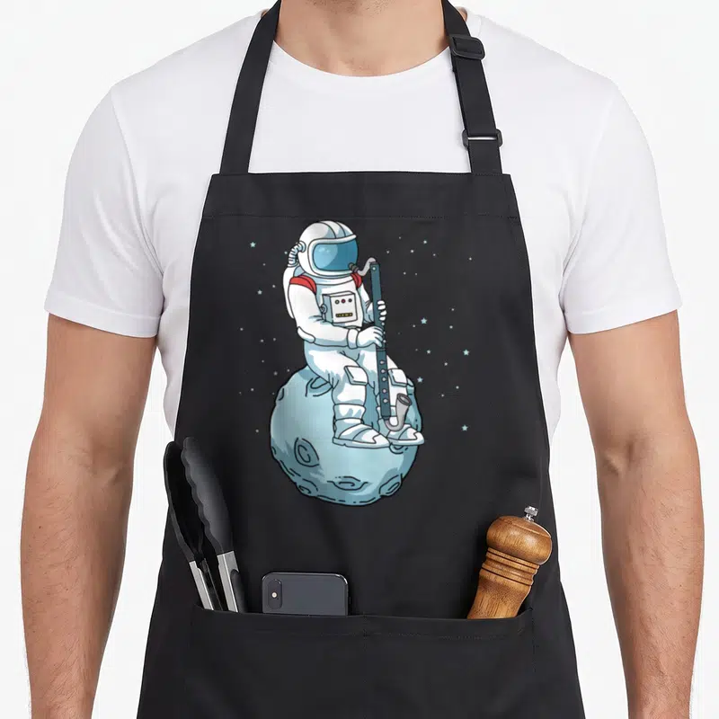 Bass clarinet apron - Cartoon astronaut in white suit sitting on blue moon holding a long dark blue didgeridoo — shown on a black cotton canvas apron against a white background