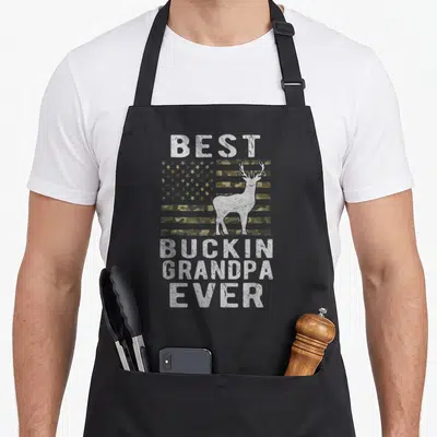 Bucking grandpa apron - Camo US flag with white deer silhouette and distressed text Best Buckin Grandpa Ever — shown on a black cotton canvas apron against a white background