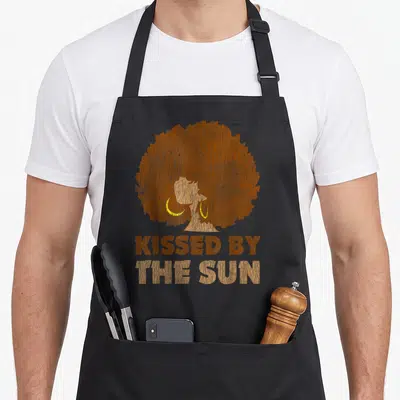 Kissed african print apron - Silhouette of a woman with a large afro and stacked text reading Kissed By The Sun — shown on a black cotton canvas apron against a white background