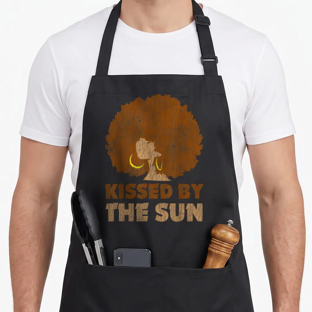 Kissed african print apron - Silhouette of a woman with a large afro and stacked text reading Kissed By The Sun — shown on a black cotton canvas apron against a white background
