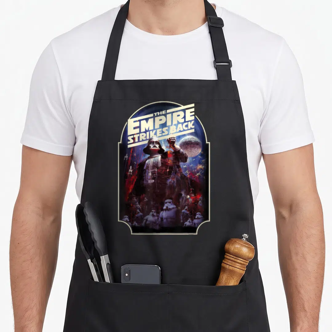 Empire novelty apron - Darth Vader and Stormtroopers inside an arched frame with The Empire Strikes Back text — shown on a black cotton canvas apron against a white background