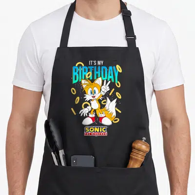 Birthday ring apron - Cartoon fox holding peace sign surrounded by gold rings under birthday text — shown on a black cotton canvas apron against a white background