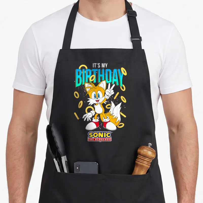 Birthday ring apron - Cartoon fox holding peace sign surrounded by gold rings under birthday text — shown on a black cotton canvas apron against a white background